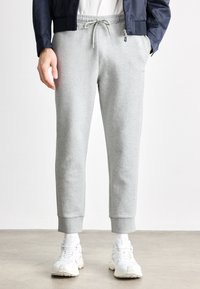 Világosszürke, puha anyagból készült melegítőnadrág rugalmas derékrésszel, húzózsinórral és bordázott mandzsettákkal. Fehér tornacipővel párosítva.