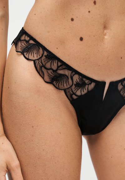 Sous-vêtements en dentelle noire avec broderie florale portés sur la hanche et le bas-ventre d'une femme avec des grains de beauté visibles et un nombril.
