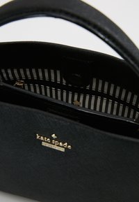 Borsa nera strutturata in materiale testurizzato con logo dorato e motivo interno a righe. Presenta un manico singolo e una tasca con cerniera.