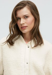 Cream-colored bomber jacket with a plush, textured exterior and smooth, contrasting front panel. Features a ribbed collar and snap closures.