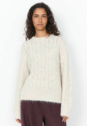 Jeune femme aux cheveux bouclés et foncés portant un pull en maille blanc et un pantalon foncé, debout devant un fond blanc uni.