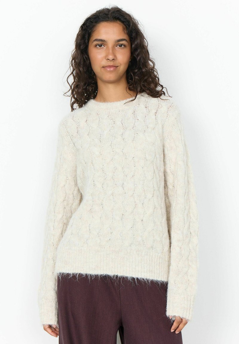 Jeune femme aux cheveux bouclés et foncés portant un pull en maille blanc et un pantalon foncé, debout devant un fond blanc uni.