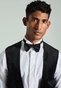 Gilet nero con finitura testurizzata sopra una camicia bianca con bottoni, caratterizzato da un papillon nero e un design elegante e moderno.