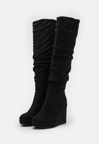 Des bottes noires au-dessus du genou en tissu doux et texturé avec un design à plis et un talon compensé, présentant une finition lisse et mate.