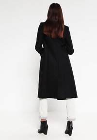 Sort lang frakke med skræddersyet design, der har en glat overflade, strømlinet silhuet og en midterste rygventil for nem bevægelse.