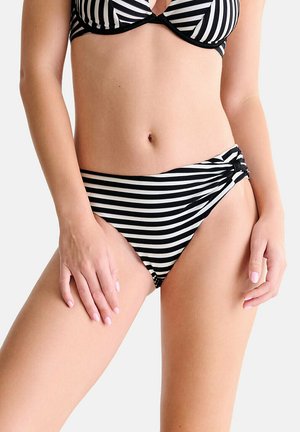 Vrouw die een zwart-witte bikinibroek draagt met zijstrikdetail en bijpassende top, staand met handen op de heupen.