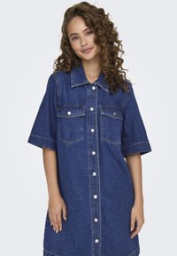 Denim shirt dress featuring short sleeves, two chest pockets, button closure, and a relaxed fit in a medium blue wash with contrasting stitching.