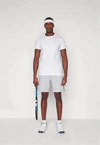 Bílé tričko s krátkým rukávem, šedé sportovní šortky, bílá čelenka, drží modrý tenisový raket; zahrnuje bílé tenisky a ponožky nad kotníky.