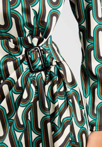 Robe à motifs avec un design graphique noir, sarcelle et orange. Présente une ceinture avec une boucle carrée et une texture plissée. Le tissu semble lisse.