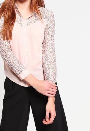 Kvinde iført en sart lyserød bluse med blomsterblondeærmer og sorte bukser med vide ben, der holder den ene arm med den modsatte hånd.
