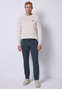 Světle béžový svetr s texturovaným vzorem, námořnické slim-fit kalhoty a šedé tenisky, přičemž svetr má detail s logem.