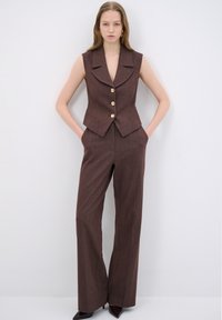 Bruine vest met een scherpe kraag en gouden knopen, gecombineerd met een wijde pantalon in een bijpassende gestructureerde stof. Model poseert met handen in de zakken.