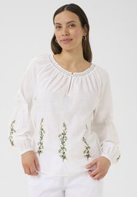 Blusa bianca in lino con maniche a sbuffo, con ricami floreali verdi e rosa lungo l'orlo e i polsini. Collo a chiave con bordo.