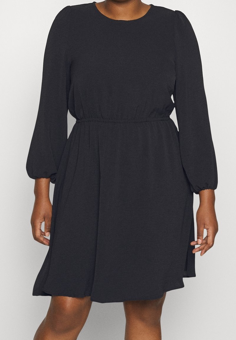 Femme portant une robe noire mi-longue aux manches longues bouffantes et à la taille élastique, debout devant un fond uni.