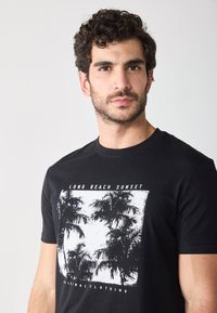 T-shirt en coton noir avec un imprimé graphique blanc de palmiers et le texte "LONG BEACH SUNSET" sur le devant. Design classique à col rond.