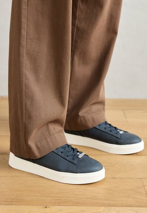 Mörkblå lädersneakers med vita gummisulor, rund tå och snörningsdesign; uppställda med lösa bruna byxor på en trägolv.