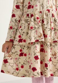 Dziecięca ręka trzyma brzeg beżowej sukienki zdobionej czerwonymi kwiatami, zielonymi liśćmi i małymi brązowymi sarnami, noszonej na różowych rajstopach o fakturze.