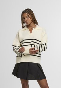 Biely pletený sveter s čiernymi horizontálnymi pruhmi, výstrih do V a rebrované manžety. Kombinovaný s čiernou plisovanou sukňou.