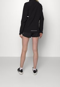Chaqueta deportiva negra con un cuello alto, textura suave y detalles reflectantes. Combinada con pantalones cortos negros y zapatillas de correr.