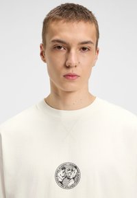 Creme sweatshirt med en rund sort grafik, der viser to boksere. Ribbet rund hals og let struktur. Rent design med minimale accenter.