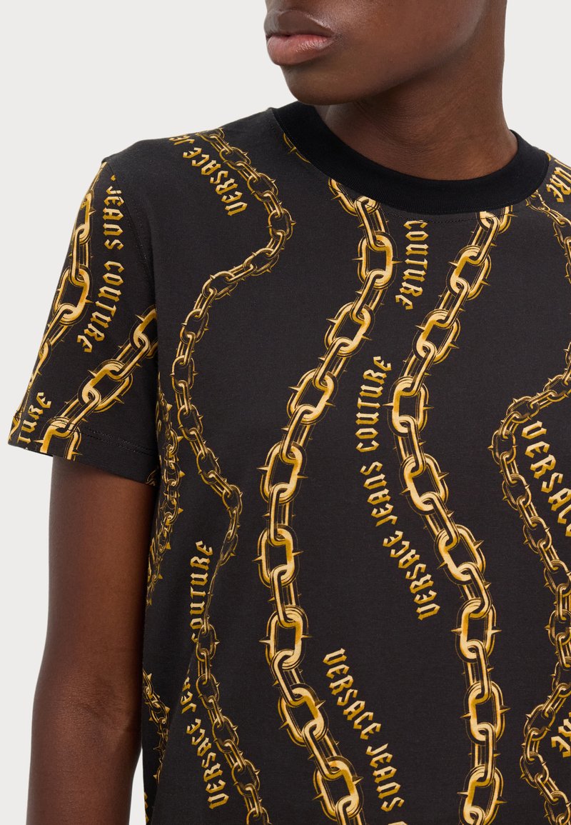 Camiseta de algodón negra con un estampado de cadenas doradas y texto que dice "Versace Jeans Couture". Diseño de manga corta y cuello redondo.