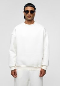 Witte crewneck sweatshirt gemaakt van zacht materiaal, met verlaagde schouders en een geribbelde onderkant. Geselecteerd met bijpassende broek en zwarte zonnebril.