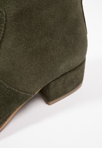 Bottine en daim olive avec un petit talon bloc, texture lisse et détails de couture subtils. Le design présente un bout arrondi.