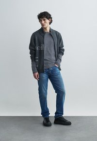 Antraciet gebreide cardigan met een gestreept grijs overhemd eronder, gecombineerd met blauwe spijkerbroek en zwarte sneakers. Simpele, zichtbare textuurdetails.