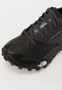 Chaussure de sport noire légère avec une tige texturée arborant des rayures jaunes, maintenue par des lacets gris, et un logo proéminent sur la semelle.