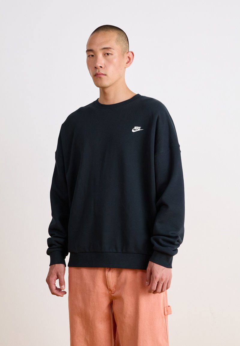 Fekete, kereknyakú pulóver, ribbelt ujjak és alj, az elülső bal mellen egy kis fehér Nike logóval. Világos őszibarack színű nadrággal viselt.