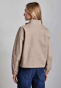 Beige, lyhyellä mallilla oleva takki, jossa on korkea kaulus, joustavat hihansuut ja sileä pinta. Yhdistetty tummansinisiin farkkuihin, joissa on hyvin istuva muotoilu.