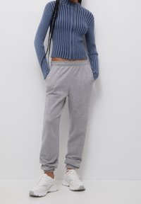 Blauw geribbelde gebreide zip-up top met een hoge kraag, gecombineerd met grijze zachte joggerbroeken en witte sneakers. Model staat tegen een effen achtergrond.