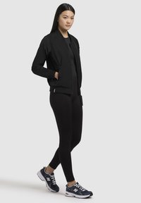 Svart bomberjacka med dragkedja, sidofickor, ribbade ärmar, bärs över svarta leggings och blå sportskor. Enkelt design, slät textur.