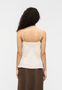 Εκρού μεταξωτό τοπ καμί με λεπτές ρυθμιζόμενες τιράντες και ελαφρώς καμπυλωτό τελείωμα, φορεμένο πάνω από καφέ φούστα. Ομαλή υφή, λιτός σχεδιασμός.