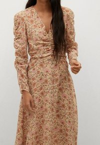 Robe florale à manches longues avec un décolleté en V, présentant une base beige clair et des motifs de fleurs roses, des détails froncés à la taille et une jupe fluide.