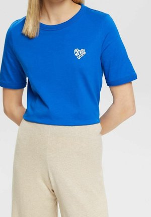 Kobieta w jasnoniebieskiej koszulce z krótkim rękawem z małym białym wzorem serca i napisem "ALL WE NEED IS FRIEND" na piersi.