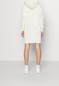 Sweatshirtklänning, krämfärgad, med huva och ribbade muddärmar, mitt på låret längd, mjukt tyg, med synliga sömnadsdetaljer och matchad med vita sneakers.