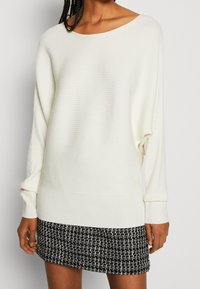 Witte geribde trui met een oversized pasvorm en een boot neckline. Gepaard met een zwart-witte textuurrrok met een ruitpatroon.