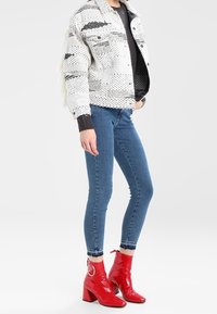 Kvinna iklädd en vit stickad jacka, mörk tröja, smala blå jeans och glänsande röda ankelboots med blockklackar mot en enkel bakgrund.