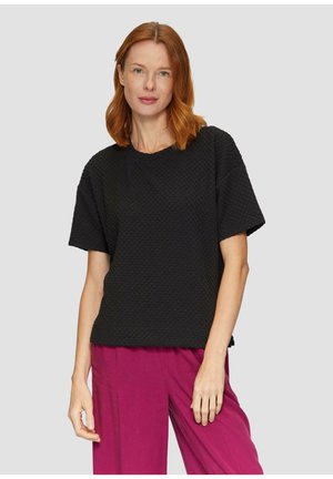 Haut noir texturé à manches courtes avec un col rond. Associé à un pantalon large rose. Modèle se tenant contre un fond gris clair.