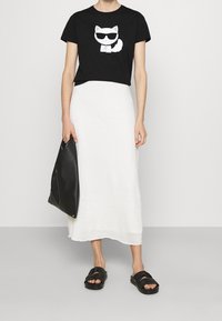 Černé croptop tričko s grafikou bílé kočky v brýlích, spárované s dlouhou bílou sukní, černými sandály a černou plážovou kabelkou.
