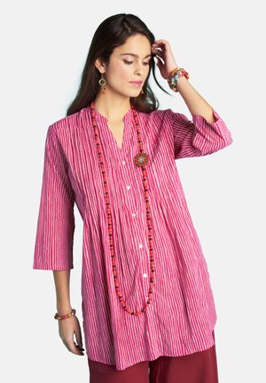 Woman wearing a pink and white striped long shirt with red pants, colorful beaded necklace, earrings, and bracelets, touching hair with one hand.