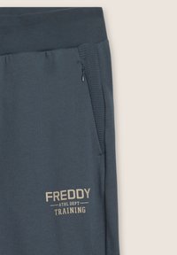 Pantaloni jogger blu scuro realizzati in tessuto morbido con tasca laterale, vita a costine e la scritta "FREDDY ATHL DEPT TRAINING" stampata in beige.