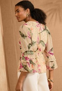Blusa floreale trasparente color crema con maniche a volant, dotata di una vita arricciata e dettagli metallici, arricchita da stampe botaniche.
