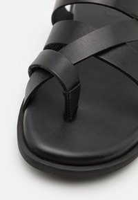 Fekete szandál sima bőr felsőrésszel, átlapolt pántokkal, lapos talppal és egyetlen lábujjpánttal. Minimalista dizájn, elegáns kivitelezéssel.