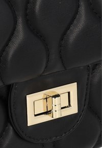 Bolso de cuero negro con textura acolchada, que presenta un cierre de hardware dorado y detalles de costura. Diseño compacto y estructurado.