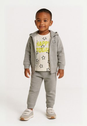 Grauer Zip-Hoodie mit Smiley-Detail, weißes T-Shirt mit "HAPPY TODAY" in leuchtendem Gelb und Sternenmuster, graue Jogginghosen und Turnschuhe.