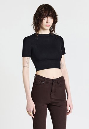 Giovane con i capelli castani bagnati e arruffati, che indossa un crop top nero aderente e pantaloni marroni scuri a vita alta, in piedi su uno sfondo bianco.