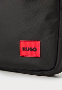 Geantă texturată neagră, având o dungă rectangulară roșie cu logo-ul „HUGO” embosat. Materialul pare durabil, cu un finisaj neted.