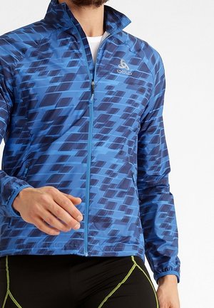 Homme portant une veste zippée bleue à motif et un pantalon de sport noir avec des surpiqûres vertes, montré du cou aux hanches.